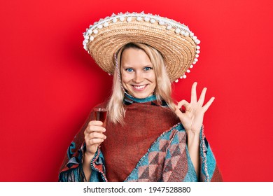 Beautiful caucasian blonde woman wearing festive mexican poncho drinking tequila shot doing ok sign with fingers, smiling friendly gesturing excellent symbol 