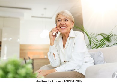 Beautiful Caucasian elderly female having phone conversation with her old friend using electronic device, discussing latest news with cute smile on her face, sitting at home
