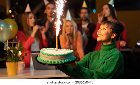 Beautiful Caucasian Female Holding A Cake As Her Co-Workers Cheer Her On In The Background Wearing Party Hats. Caucasian Office Workers Throwing A Surprise Birthday Party For Their Co-Worker.