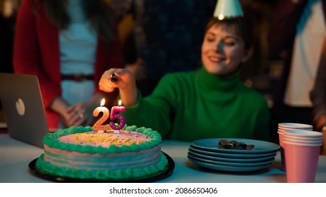 Beautiful Caucasian Female Sitting Behind a Desk Wearing a Green Sweater and a Party Hat With a Birthday Cake In Front Of Her, Holding a Lighter, Lighting Up The Candles. Surprise Birthday Party. - Powered by Shutterstock