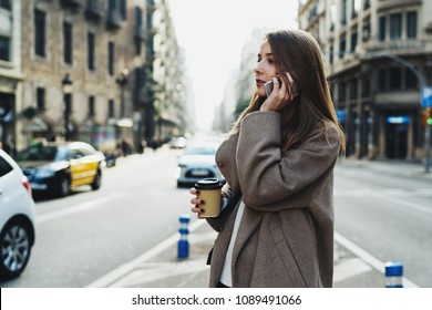 Beautiful caucasian female is talking by a smartphone while standing on the street with a take away coffee. Charming lady in stylish coat looking aside while chatting with a friend by a mobile phone.