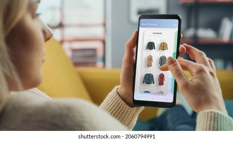 Beautiful Caucasian Female Is Using Smartphone With Clothing Online Web Store To Choose And Buy Clothes From New Collection. Female Surfing The Net And Lying On Couch Sofa At Home Living Room.