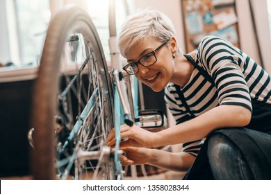 Beautiful Caucasian female worker with short blonde hair and eyeglasses crouching and repairing bicycle. Bike workshop interior.