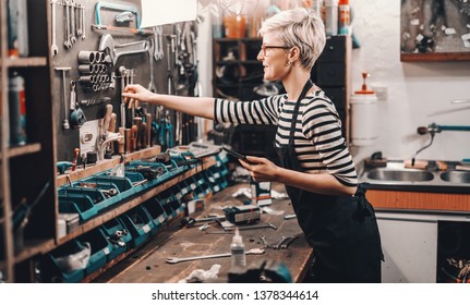 Beautiful Caucasian female worker with short blonde hair and eyeglasses taking tool fromm wall to repair bicycle. Bike workshop interior. - Powered by Shutterstock