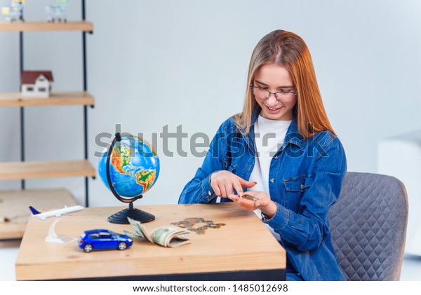 Beautiful caucasian girl\
counts her savings to foreign vacation. Young woman sits at table\
with glass jar, money, globe, model of air plane and car. Close up,\
selective focus.