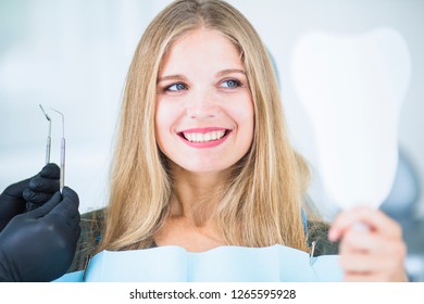 Beautiful Caucasian Girl In Dentist Blue Bib Sit In The Dentist Chair With Mirror In Hands, Looking At Herself, Smiling