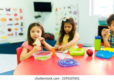 Beautiful Caucasian Girl Eating A Sandwich With Preschool Friends And Enjoying The Lunch Break In The Classroom