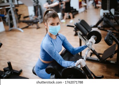 A Beautiful Caucasian Girl With A Mask And Gloves Stands And Exercises With Weights In The Gym. COVID - 19 Virus Protection