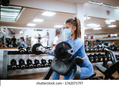 A Beautiful Caucasian Girl With A Mask And Gloves Stands And Exercises With Weights In The Gym. COVID - 19 Virus Protection