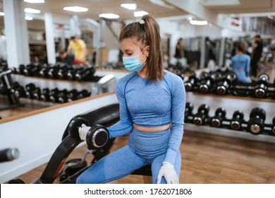 A Beautiful Caucasian Girl With A Mask And Gloves Stands And Exercises With Weights In The Gym. COVID - 19 Virus Protection