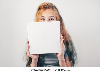 Beautiful caucasian girl with mockup poster, grey wall on background. Girl holding in her hands square blank canvas board.