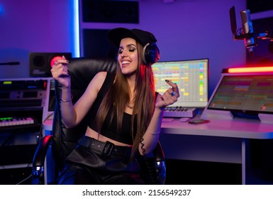 Beautiful caucasian girl in stylish clothes works in a recording studio with neon light and goes crazy in a cap and glasses