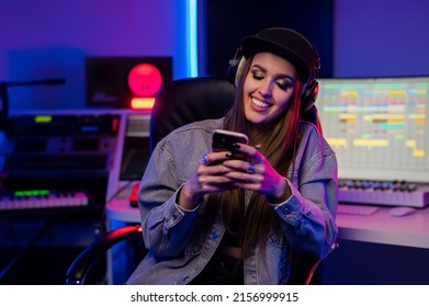 Beautiful caucasian girl in stylish clothes in headphones looks at the phone and smiles in the recording studio with neon light - Powered by Shutterstock