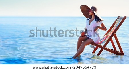  Beautiful caucasian girl in sunhat and white tunic sitting on the beach chair at seaside while holds cocktail. Looking in sideways. Summer vacation. Skin care