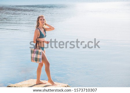 Beautiful caucasian girl in a swimsuit with a modern beach bag on the seashore smiles and looks at the camera.