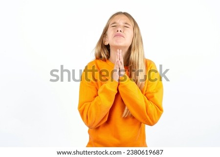 beautiful caucasian kid girl orange sweater using smartphone begging and praying with hands together with hope expression on face very emotional and worried. Please God