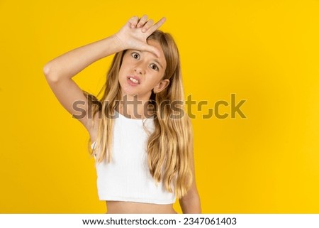beautiful caucasian kid girl wearing white T-shirt making fun of people with fingers on forehead doing loser gesture mocking and insulting.