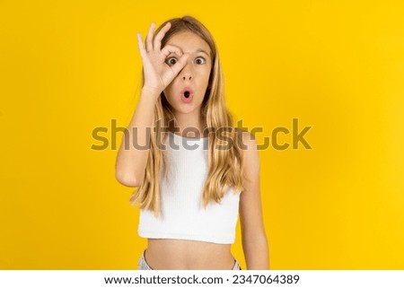 beautiful caucasian kid girl wearing white T-shirt doing ok gesture shocked with surprised face, eye looking through fingers. Unbelieving expression.