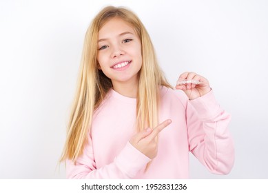 beautiful caucasian little girl wearing pink sweater over white background holding an invisible aligner and pointing at it. Dental healthcare and confidence concept.