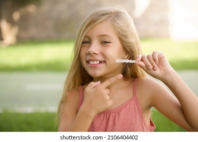 Beautiful Caucasian Little Kid Girl Wearing Dress Standing Outdoors  Holding An Invisible Aligner And Pointing At It. Dental Healthcare And Confidence Concept.