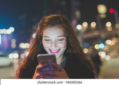 Beautiful Caucasian Long Brown Hair Woman Using Smart Phone Hand Hold Outdoor In The City Night, Smiling, Face Illuminated Screenlight - Social Network, Technology, Communication Concept