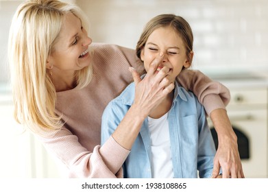 Beautiful Caucasian Middle Aged Blonde Woman And Preschooler Girl In The Kitchen. Grandma Hugging Granddaughter, Touching Her Nose Finger With Flour, They Have Fun Together At Home