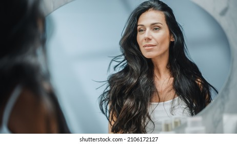 Beautiful Caucasian Middle Aged Woman Looks Into Bathroom Mirror, Enjoys Her Looks. Concept For Happiness, Wellbeing, Natural Beauty And Organic Skin Care Products