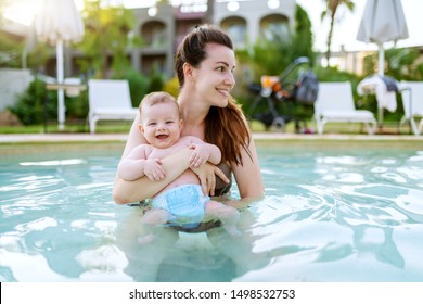 Beautiful Caucasian mother standing in swimming pool and holding her 6 months old son. Baby looking at camera and smiling.