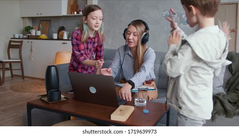 Beautiful Caucasian Mother Trying To Work Home Online Using Laptop, Distracted By Two Teenage Children Needing Attention