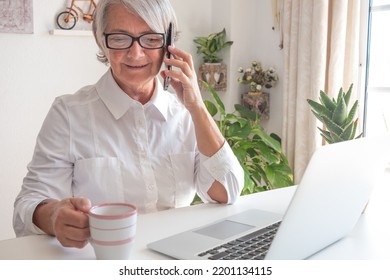 Beautiful Caucasian Senior Woman In White Shirt Using Laptop While Working Indoors Holding Mobile Phone In Hand, Smiling Lady Talking At Cellphone