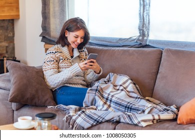 Beautiful Caucasian Smiling Woman Sitting On Cozy Couch And Typing On Cell Phone.