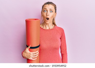 Beautiful caucasian sports woman holding yoga mat scared and amazed with open mouth for surprise, disbelief face 
