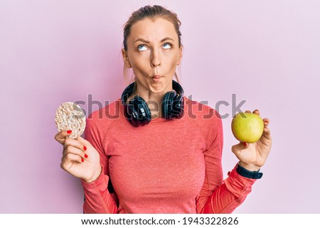 Beautiful caucasian sports woman holding green apple and rice crackers making fish face with mouth and squinting eyes, crazy and comical. 