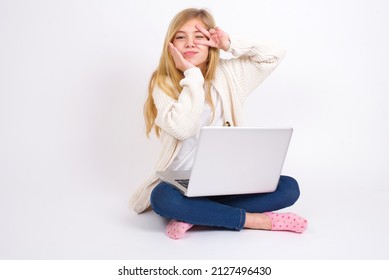 Beautiful caucasian teen girl sitting with laptop in lotus position on white background  making v-sign near eyes. Leisure lifestyle people person celebrate flirt coquettish concept.