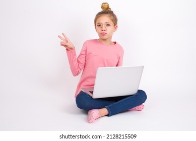 Beautiful caucasian teen girl sitting with laptop in lotus position on white background makes peace gesture keeps lips folded shows v sign. Body language concept