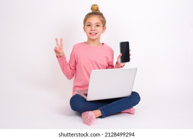 Beautiful caucasian teen girl sitting with laptop in lotus position on white background holding modern device showing v-sign