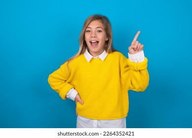 beautiful caucasian teen girl wearing yellow sweater over blue wall holding finger up having idea and posing