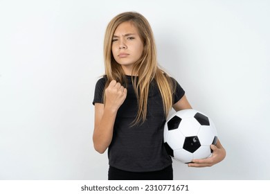 beautiful caucasian teen girl wearing sportswear holding a football ball over white wall shows fist has annoyed face expression going to revenge or threaten someone makes serious look.  - Shutterstock ID 2310771671