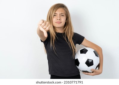 beautiful caucasian teen girl wearing sportswear holding a football ball over white wall looking unhappy and angry showing rejection and negative with thumbs down gesture. Bad expression. - Shutterstock ID 2310771791