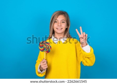 beautiful caucasian teen girl wearing yellow sweater over blue wall showing and pointing up with fingers number two while smiling confident and happy.