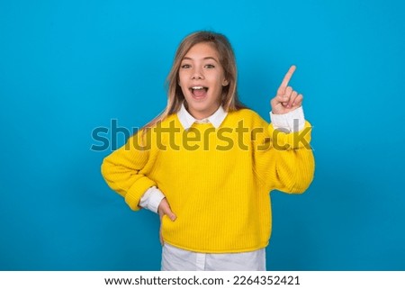 beautiful caucasian teen girl wearing yellow sweater over blue wall holding finger up having idea and posing