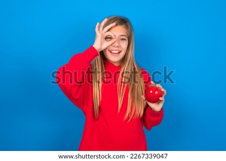 Beautiful caucasian teen girl wearing red sweatshirt over blue background eating apples with happy face smiling doing ok sign with hand on eye looking through finger.