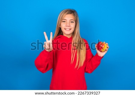 Beautiful caucasian teen girl wearing red sweatshirt over blue background eating apples showing and pointing up with fingers number two while smiling confident and happy.