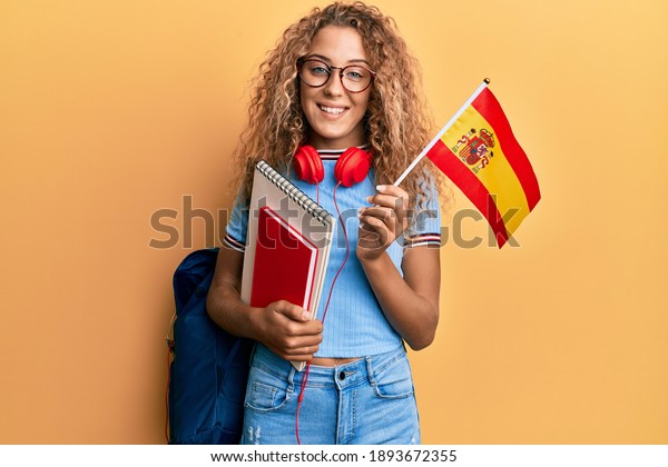Beautiful caucasian teenager girl exchange student\
holding spanish flag smiling with a happy and cool smile on face.\
showing teeth. 