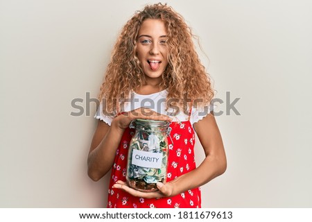 Beautiful caucasian teenager girl holding charity jar with money sticking tongue out happy with funny expression. 