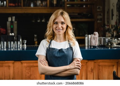 Beautiful caucasian waitress small business owner barista bartender standing in blue apron looking at the camera with arms crossed at the bar counter in restaurant.