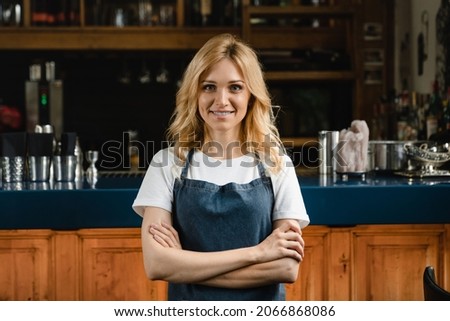 Beautiful caucasian waitress small business owner barista bartender standing in blue apron looking at the camera with arms crossed at the bar counter in restaurant.