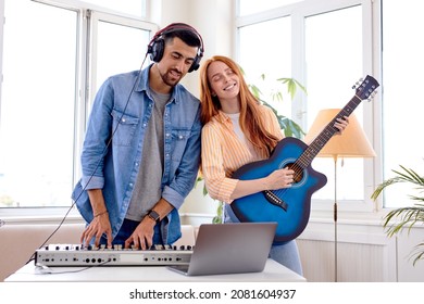 Beautiful Caucasian Woman Artist Enjoying The Process Of Playing Guitar While Handsome Bearded Guy Playing Piano, Duet In Bright Studio Room, Using Laptop. Music, People Lifestyle Concept