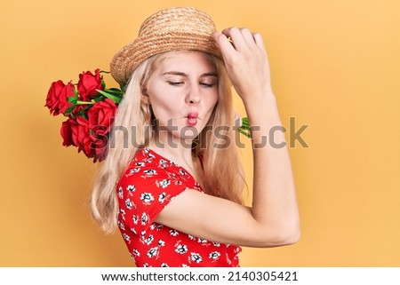 Beautiful caucasian woman with blond hair holding bouquet of red roses making fish face with mouth and squinting eyes, crazy and comical. 