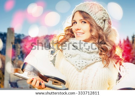 Beautiful caucasian woman going to ice skating outdoor. She dressed in white winter pullover and warm hat. Holding skates shoes. Healthy lifestyle and sport concept at olympic stadium at nature.